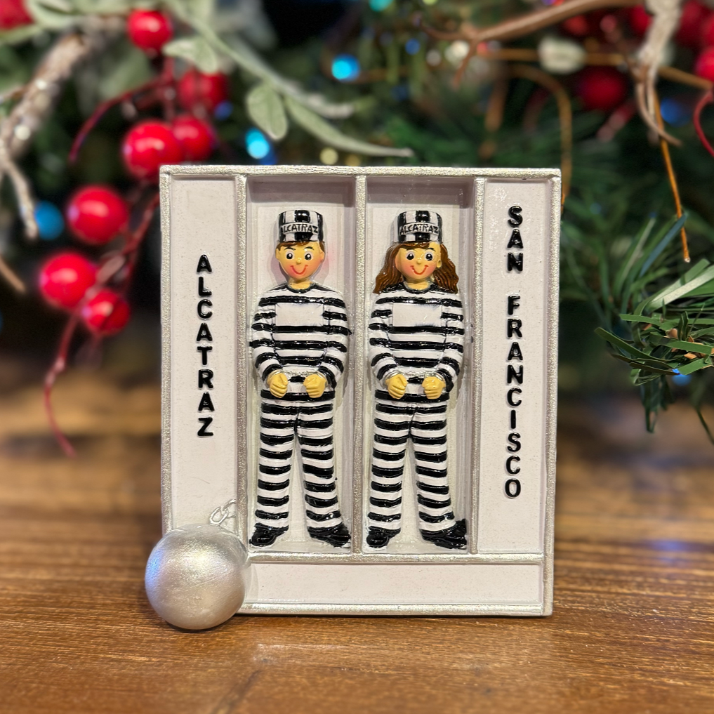 Two small figures in striped prison uniforms in a jail cell labeled 'Alcatraz' and 'San Francisco', with Christmas decorations in the background.