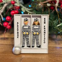 Two small figures in striped prison uniforms in a jail cell labeled 'Alcatraz' and 'San Francisco', with Christmas decorations in the background.